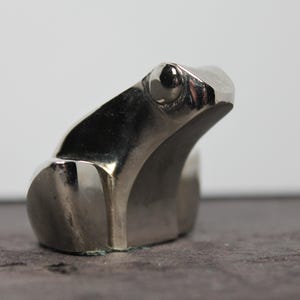 May include: A small, polished silver-toned metal frog figurine with a smooth, reflective surface. The frog is depicted in a seated, upright position with its head turned slightly to the side, showcasing a minimalist, solid form and rounded features.