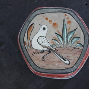 May include: A hand-painted, hexagonal ceramic tile featuring a white bird with black details, perched amidst stylized plants. The bird is set against a tan background, framed by a gray border with red and black accents. Decorative art.