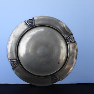 May include: A silver-colored plate with a wide, curved rim and a central, slightly concave area. The rim features decorative, fan-shaped accents at intervals. The plate is set against a light blue background.
