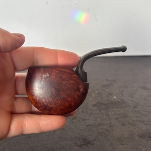 May include: A brown, curved pipe with a dark stem. The pipe has a rounded bowl with a wood grain pattern. The stem is dark and curved, extending from the bowl. The pipe is held in a hand.