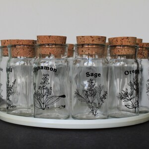 May include: A clear glass spice rack with cork stoppers. Each jar is labeled with a spice name in black lettering and features a botanical illustration. The rack is on a white, circular base.