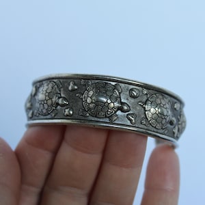 May include: A silver-toned metal cuff bracelet featuring a repeating embossed pattern of turtles. The surface has a darkened, oxidized finish in the recessed areas, highlighting the detailed shell textures and shapes of the turtles along the band.