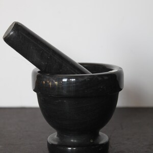 May include: A black mortar and pestle set. The mortar is a rounded bowl with a wide rim, and the pestle is a cylindrical shape. Both pieces have a polished, glossy finish. The set is designed for grinding herbs and spices.