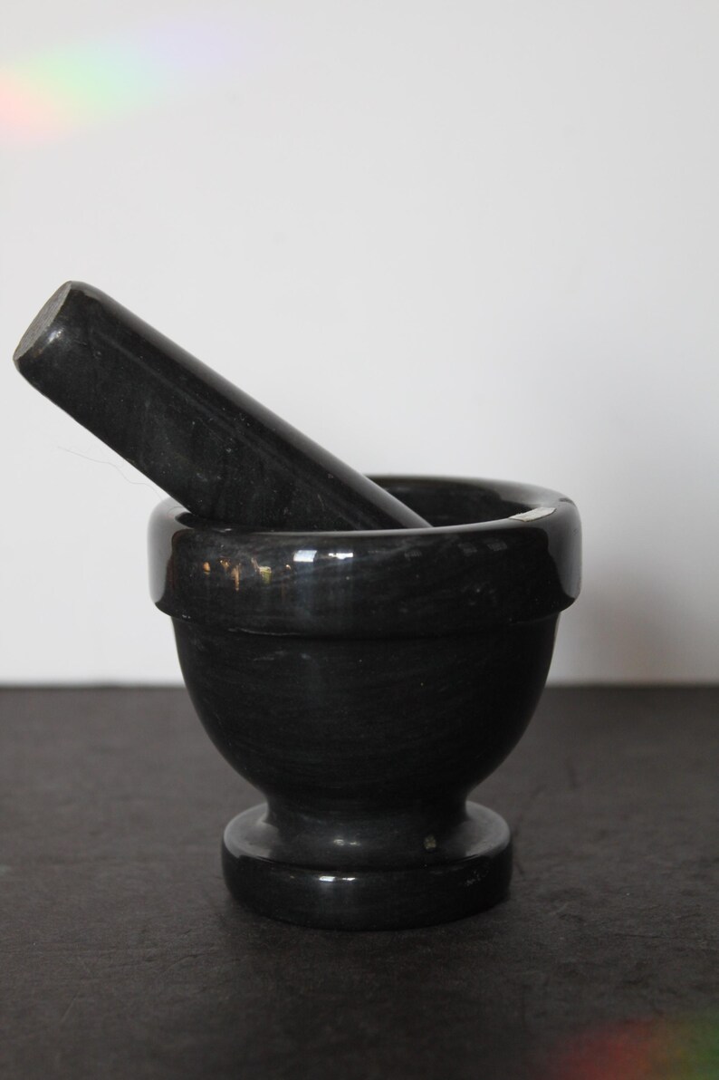 May include: A black mortar and pestle set. The mortar is a rounded bowl with a wide rim, and the pestle is a cylindrical shape. Both pieces have a polished, glossy finish. The set is designed for grinding herbs and spices.
