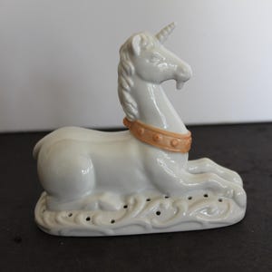 May include: White porcelain unicorn figurine with a gold and orange collar. The unicorn is lying down with its head turned to the side. The base of the figurine is decorated with a floral design.