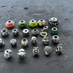 May include: A collection of various beads and charms, including silver, glass, and gemstone designs. The beads feature diverse shapes, colors, and patterns, such as stripes, polka dots, and floral motifs. Some charms have sparkling accents.