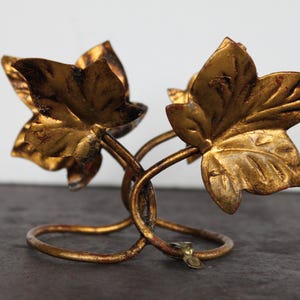May include: A decorative, golden-colored metal leaf-shaped candle holder. The candle holder features two stylized leaves and a looped base. The leaves and base have an aged, distressed finish, adding to its vintage appeal. The candle holder is designed to hold two candles.