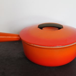 May include: A bright orange enameled cast iron saucepan with a matching lid and a long handle. The pot features a gradient color transition from a deep red-orange at the top to a lighter, vibrant orange at the base, topped with a dark brown handle.