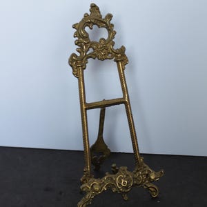 May include: A brass easel stand featuring an ornate, scrollwork top and a matching decorative base. The frame consists of two vertical supports with a textured, rope-like pattern and a horizontal crossbar, designed to hold items for display.