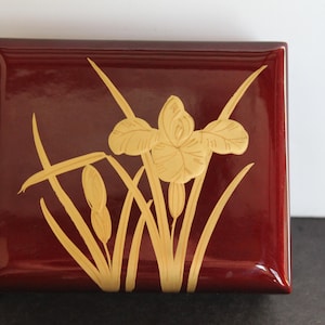 May include: A rectangular, burgundy-colored box with a glossy finish. The lid features a carved, light-yellow iris flower and cattails design. The box is likely used for storing small items.