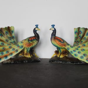 May include: Two peacock figurines with outstretched tail feathers. The birds have blue crests, black bodies, and colorful plumage in shades of green, yellow, and orange. They are perched on a dark base.