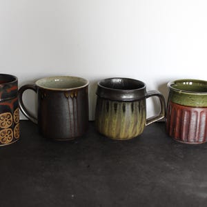May include: Four ceramic mugs in a row, each with a unique design. The mugs feature a variety of colors, including brown, green, and red. One mug has a floral pattern, while another has vertical ridges.