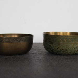May include: Two decorative metal bowls. One is bronze-colored with a patterned band near the rim. The other is green with an etched floral design. Both bowls have a gold interior and sit on a dark surface.