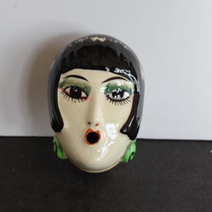 May include: A ceramic figurine of a woman's head with black hair, green earrings, and a surprised expression. The figurine is painted with black, white, green, and orange paint.