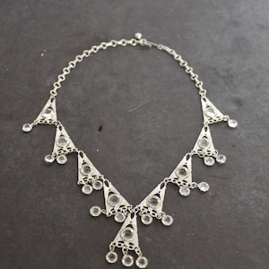 May include: A silver necklace with a geometric design and clear crystal accents. The necklace has a chain link style and features a series of triangular shapes with dangling crystals.