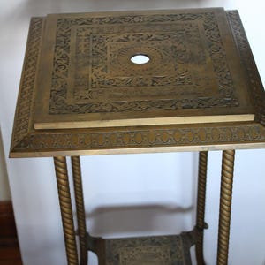 May include: A square brass plant stand featuring a top and lower shelf with intricate engraved patterns and a central circular hole. The stand is supported by four twisted brass legs that connect to a decorative base with ornate metal detailing.