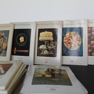 May include: A collection of vintage cookbooks titled "Recipes of the World." The covers feature illustrations of various cuisines, including Scandinavian, Japanese, and Russian cooking. The books are spiral-bound and stacked on a dark surface.