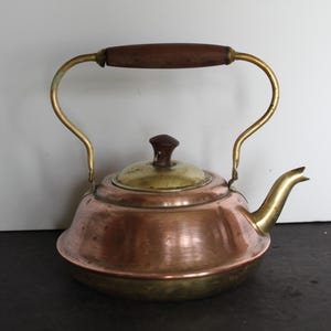 May include: A vintage copper and brass tea kettle with a wooden handle. The kettle has a rounded body, a brass spout, and a brass handle that arches over the top. The lid is brass with a small wooden knob.