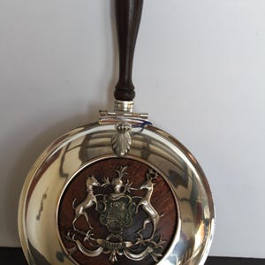 May include: A silver-plated serving dish with a dark wooden handle and a circular design. The center features a wooden inlay with a coat of arms and two deer. The handle is dark brown.