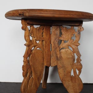 May include: A small, round wooden stool with intricate floral carvings on the legs and supports. The stool is made of light brown wood and has a simple, circular top. The legs are carved with detailed leaf and vine patterns.