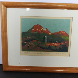 May include: A framed print featuring a landscape illustration of red mountains under a blue sky, with a central structure and small buildings. The print includes handwritten text at the bottom reading "Ghost Town, Red Mountain Colorado" and a signature.