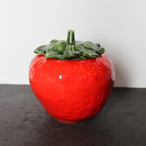 May include: A ceramic strawberry-shaped jar with a glossy red finish. The jar has a green leafy top and a small green stem. The surface of the jar is textured with small, round indentations, resembling a real strawberry.