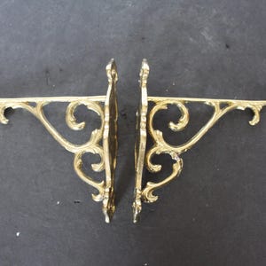 May include: Two ornate, gold-colored metal shelf brackets. Each bracket features a decorative, scrolled design and a horizontal arm for supporting a shelf. The brackets are displayed against a dark background.