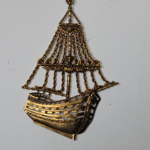 May include: A gold-colored ship pendant necklace. The pendant is a detailed representation of a sailing ship, with intricate chain detailing for the sails and rigging. The necklace hangs from a delicate gold chain, creating a vintage aesthetic.