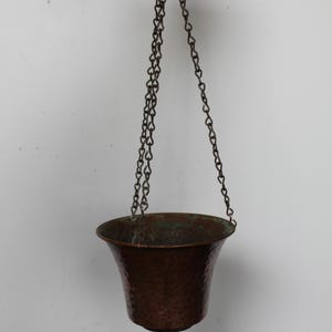 May include: A hammered copper hanging planter with a flared rim and a weathered green patina on the interior. The vessel is suspended by three thin metal chains that connect to a single circular ring at the top for easy mounting and display.