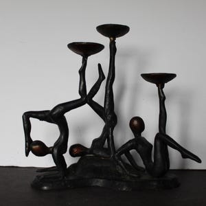 May include: A bronze sculpture of stylized human figures holding up shallow bowls. The figures are in various poses, some upside down, creating a dynamic and artistic candle holder. The sculpture has a dark brown finish.