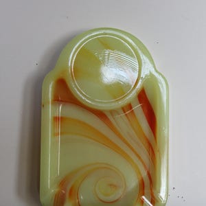 May include: A rectangular, light green glass tray with a rounded top and a circular indentation. Swirling orange and yellow patterns decorate the surface, creating a marbled effect. The tray has a glossy finish.
