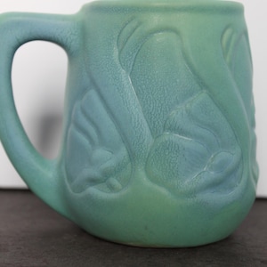 May include: A ceramic mug with a matte, textured finish in shades of teal and seafoam green. The exterior features a raised, embossed floral pattern with curved, organic shapes wrapping around the body. It includes a sturdy, curved handle.