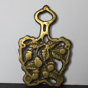 May include: A vintage brass trivet with a handle, featuring a detailed design of strawberries and leaves. The trivet is approximately 6 inches long and 5 inches wide, with a golden hue.