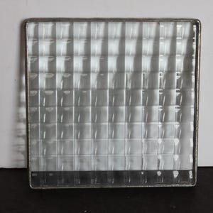 May include: A square, clear glass block with a grid pattern, framed by a thin, silver-colored metal. The glass has a textured surface, creating a visual effect of numerous small squares. The block is set against a white and black background.