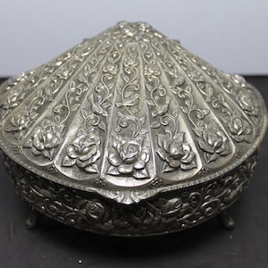May include: A silver-toned metal trinket box shaped like a seashell, featuring an intricate embossed pattern of blooming roses, scrolling vines, and small birds on the lid and sides, supported by four small, ornate metal feet.