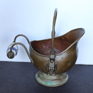 May include: A copper coal scuttle with a rounded body, a pedestal base, and a vertical handle. The piece features a lion head mount where the handle attaches and a secondary handle with a blue and white patterned ceramic grip on the side.