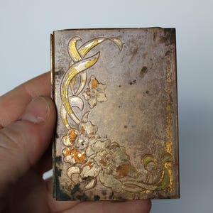 May include: A small, rectangular antique metal box with a floral design. The box is silver with gold accents and a detailed floral pattern in white, orange, and green. The box is held in a hand.