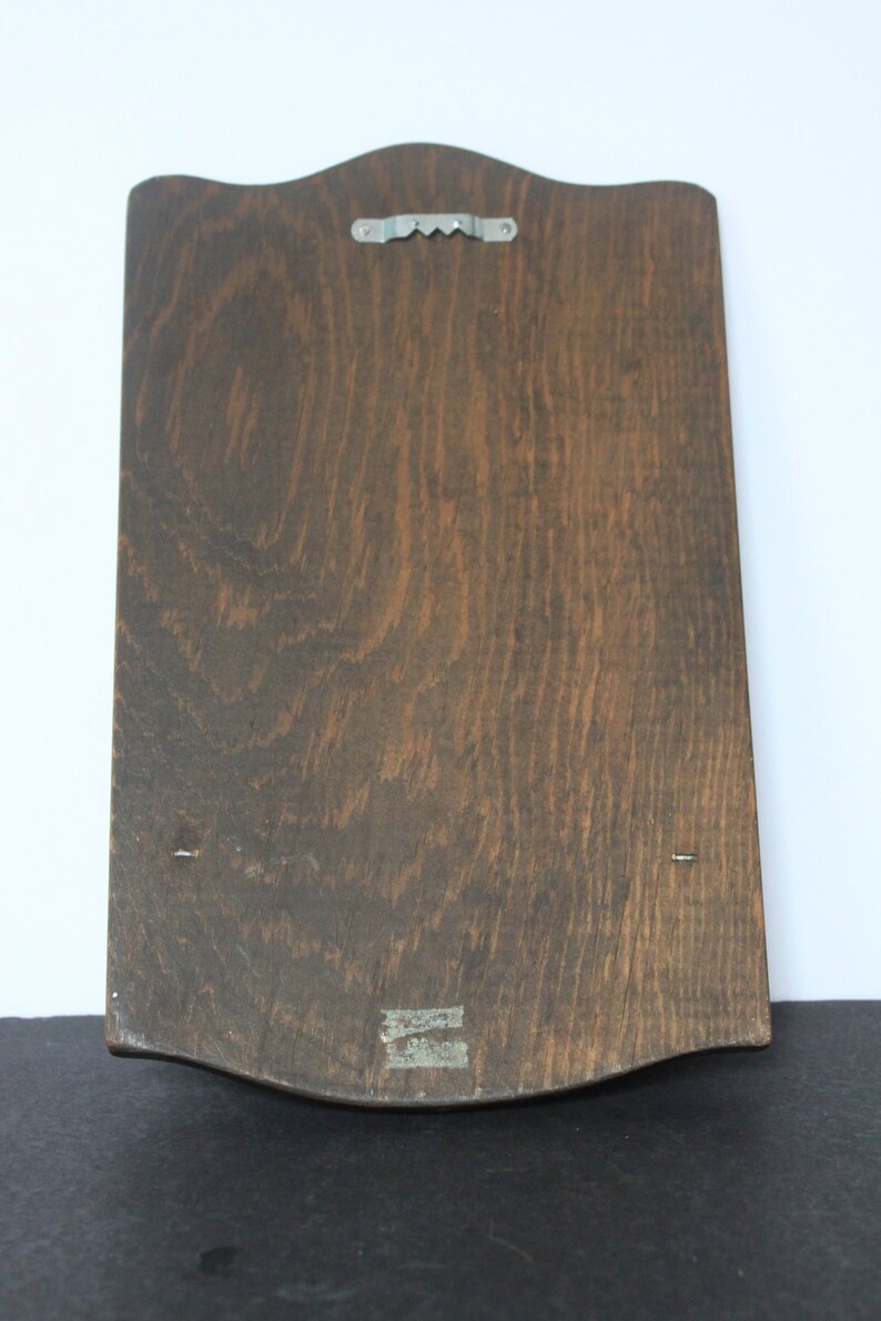 May include: A dark brown wooden plaque with a scalloped edge and a metal hook on the back. The plaque has two small nails on the bottom edge.