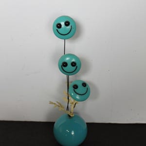 May include: A whimsical teal-colored decorative piece featuring three smiley face ornaments suspended above a teal vase. Each smiley face has black eyes and a curved smile. The vase has a round shape and is adorned with small, dried sprigs.