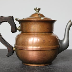 May include: A copper teapot with a wooden handle and a silver spout. The teapot has a lid with a small chain attached to the knob.