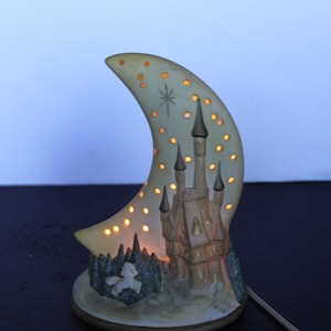 May include: A ceramic night light shaped like a crescent moon with a castle and a unicorn scene. The moon has cut-out star shapes that glow when lit. The castle has multiple spires and is painted in muted tones.