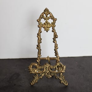 May include: A small, ornate brass easel with a decorative, openwork design. The easel features a detailed floral and scroll pattern, with a hinged back support. The overall height is approximately 6 inches.