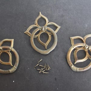 May include: Three antique brass door pulls with a decorative floral design and a ring handle. The pulls are arranged in a row on a dark gray surface.