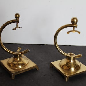 May include: Two brass candle holders with a unique design. Each holder features a square base, a central circular element, and a curved arm with a small, decorative piece at the top. The brass has a polished, golden finish.