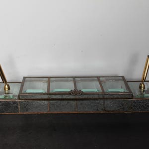 May include: A vintage desk organizer with a mirrored finish and glass compartments. It features two gold-colored pen holders and a central section with four compartments. The organizer has decorative feet and a detailed clasp.