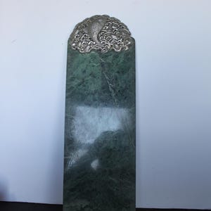 May include: A rectangular green marble slab featuring a polished surface with natural veining. The top edge is adorned with a silver-toned metal decorative piece depicting a fish swimming amidst stylized waves and swirling water patterns.