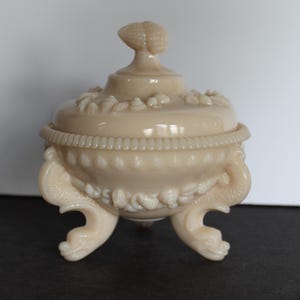 May include: A vintage, cream-colored glass candy dish with a seashell design. The dish has three legs shaped like seahorses and a lid with a seashell finial.