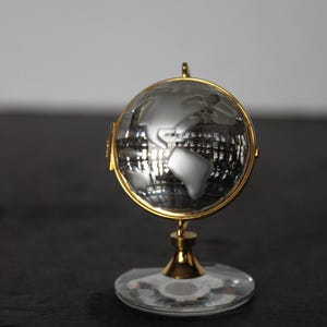 May include: A decorative globe with a silver world map encased in a clear glass sphere. The globe is held by a gold-colored metal frame and stand, with a clear, circular base. The overall design is elegant and detailed.