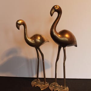 May include: Two brass flamingo sculptures. The flamingos have long, slender legs and curved necks. The sculptures are standing on leaf-shaped bases. The brass has a warm, golden hue, and the sculptures are set against a neutral background.
