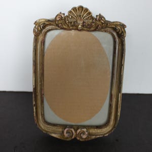 May include: Ornate, antique-style picture frame with a gold finish. The frame has a rectangular shape with a decorative shell-like design at the top and scroll details at the bottom. The frame holds a circular piece of tan paper.